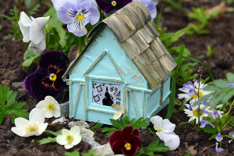 A samll blue fairy house with tiny lacy curtains is nestled in pansies in the woods. 